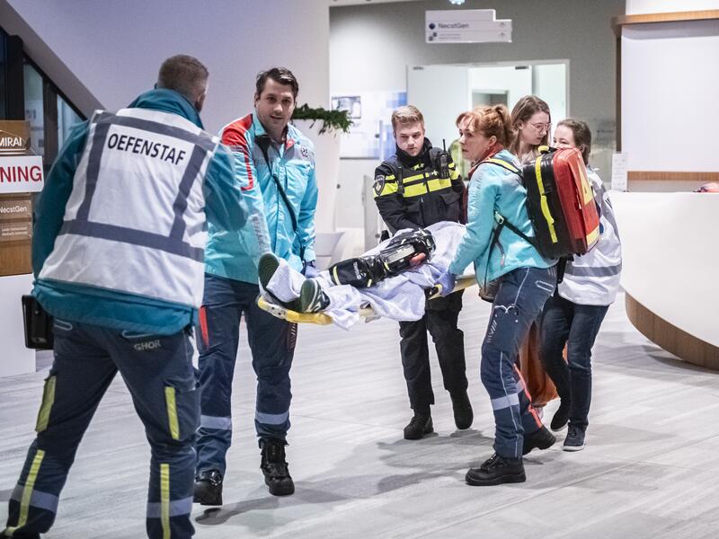 Een gewonde wordt op een brancard het pand uit gedragen door mensen van ambulance en politie. Iemand van de oefenstaf kijkt toe