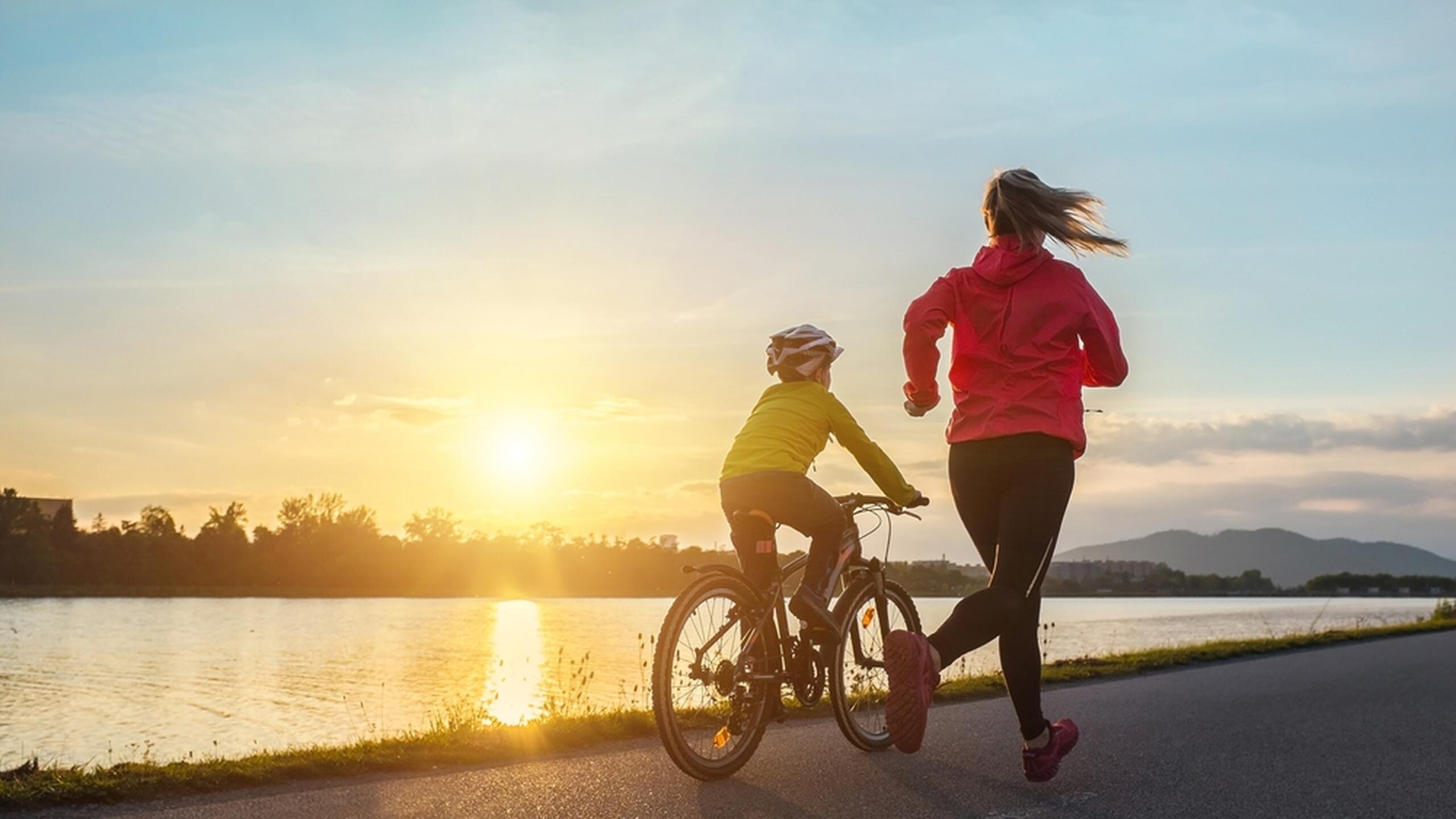 vrouw in rood jack loopt hard, kind met helm fiets er naast, op achtergrond meer en ondergaande zon