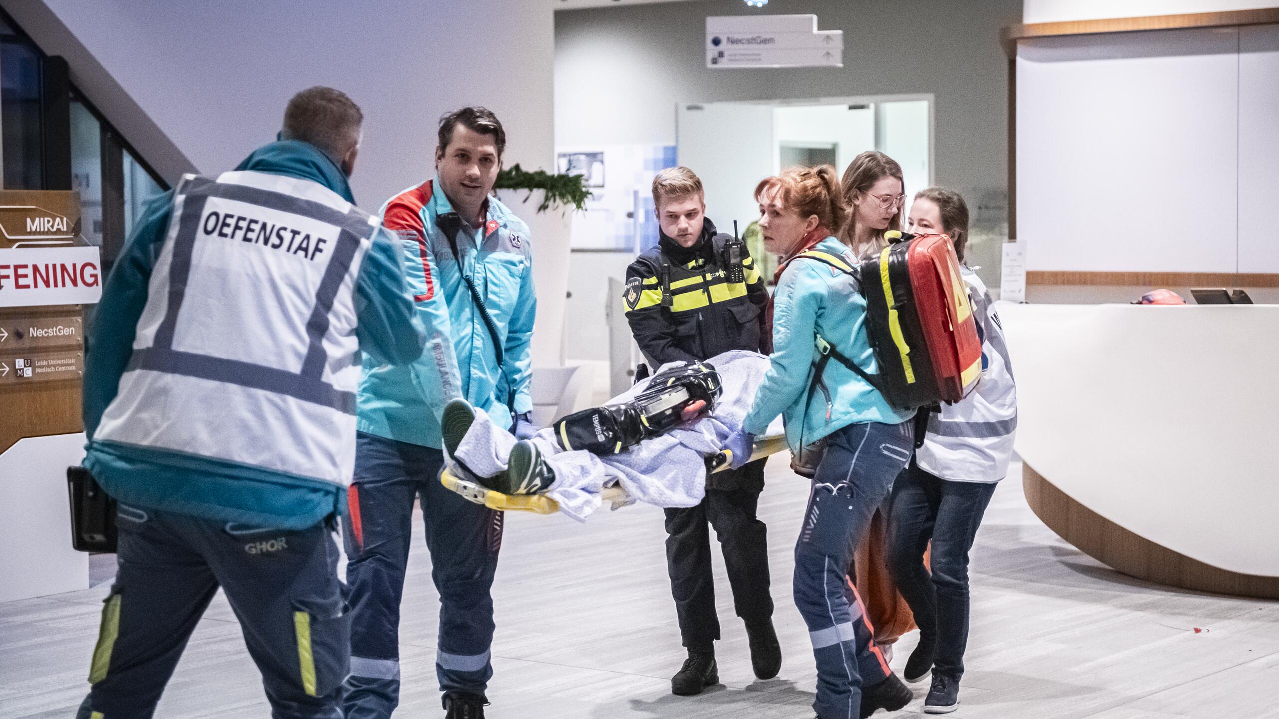 Een gewonde wordt op een brancard het pand uit gedragen door mensen van ambulance en politie. Iemand van de oefenstaf kijkt toe