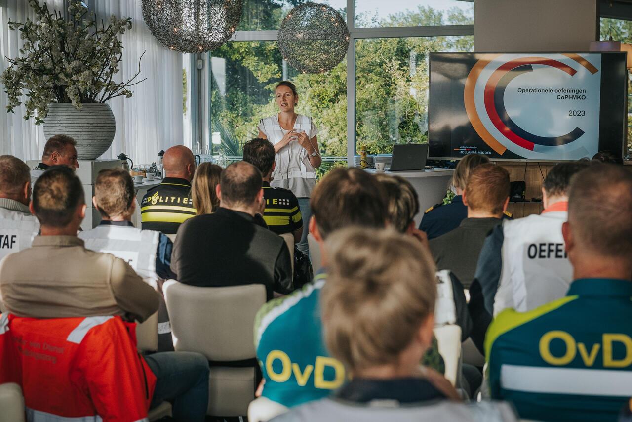 Zaal vol mensen die meedoen aan oefening, op rug gezien, voor zaal staat vrouw te praten