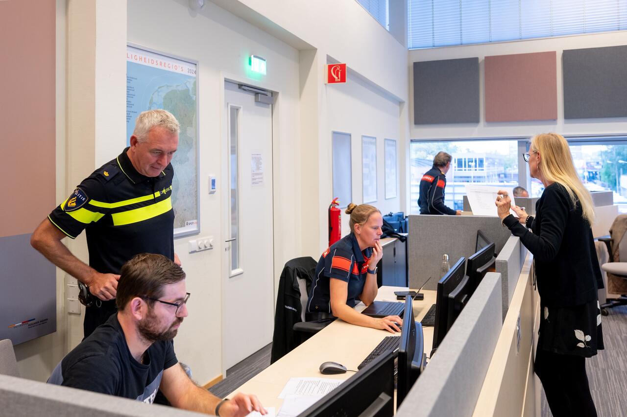 Twee mensen waarvan een in brandweeruniform achter bureau kijkend op beeldscherm. Twee mensen, waaronder een in politieuniform, kijken toe