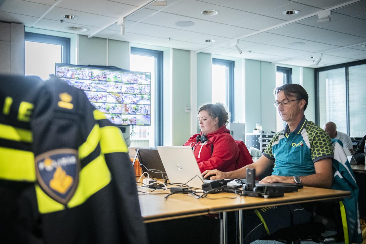 Officier van Dienst Geneeskundig geconcentreerd aan het werk in ruimte Veiligheidsteam, naast hem collega van Rode Kruis, op voorgrond jas politieuniform zichtbaar