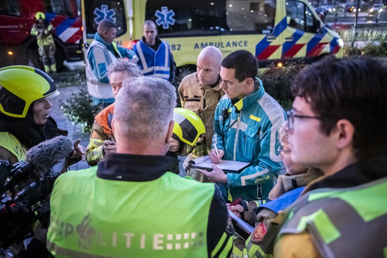 Mensen van onder andere politie, brandweer en GHOR overleggen met elkaar, buiten, op achtergrond ambulance zichtbaar