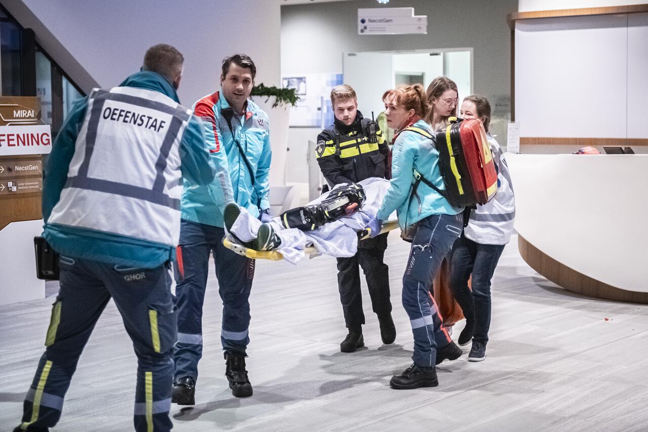 Een gewonde wordt op een brancard het pand uit gedragen door mensen van ambulance en politie. Iemand van de oefenstaf kijkt toe